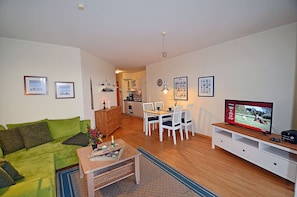 Ein Wohnzimmer mit grünem Sofa, einem Holztisch, einem Essbereich mit weißem Tisch und Stühlen, einer Küche im Hintergrund und einem Fernseher auf einem Schrank.