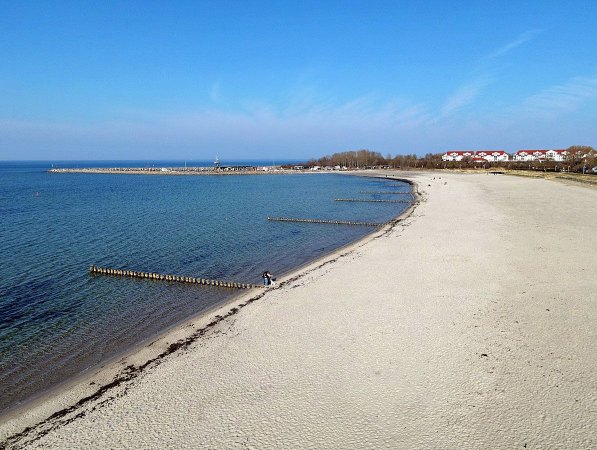 Una playa con cielo despejado, un muelle de madera y edificios al fondo.