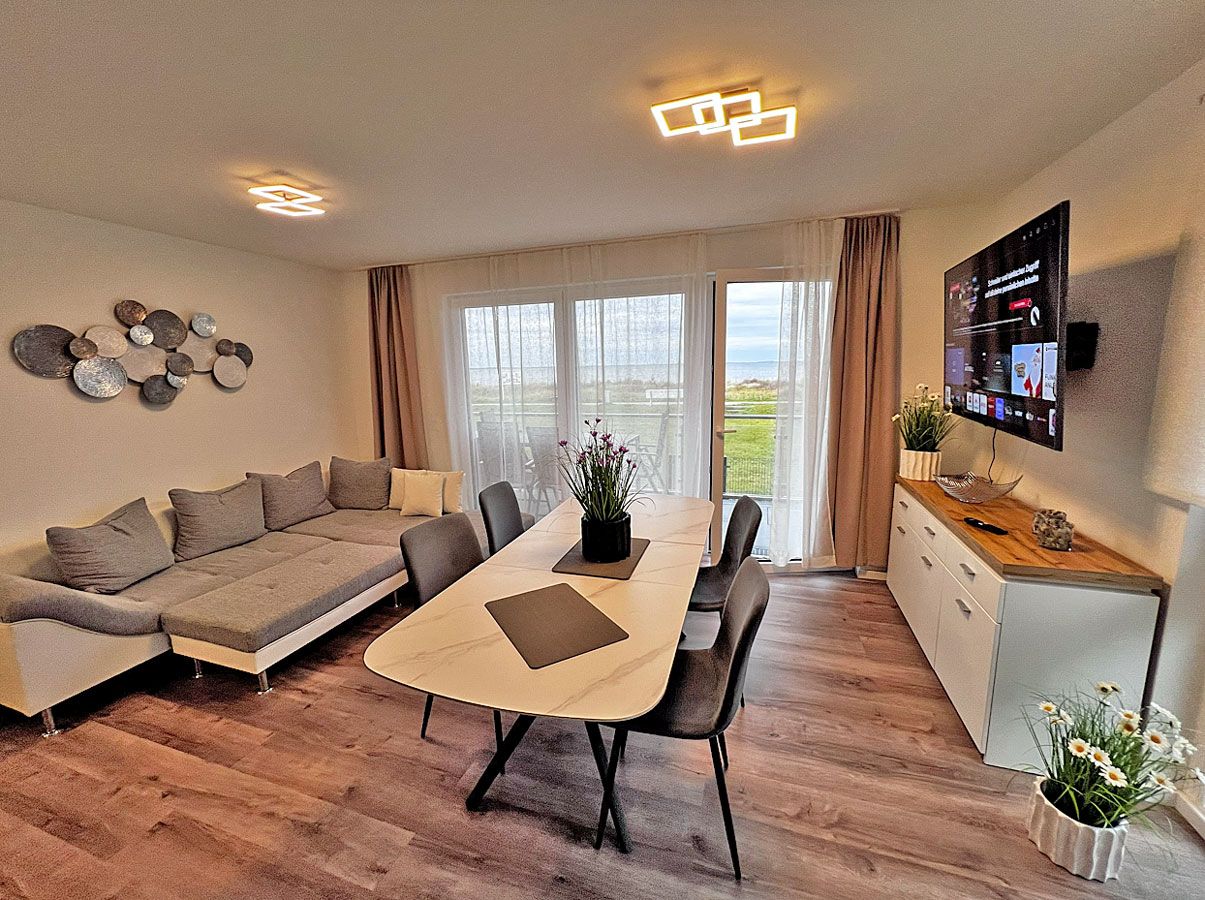 A modern living room with a sofa, dining table, chairs, a flat-screen TV, and a kitchen area with a refrigerator and cabinets.