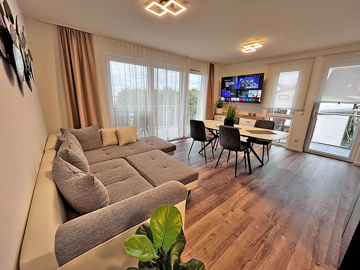 A modern living room with a sectional sofa, a dining area with a table and chairs, and a flat-screen TV mounted on the wall.