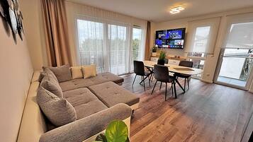 A modern living room with a sectional sofa, a dining area with a table and chairs, and a flat-screen TV mounted on the wall.