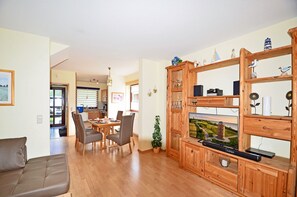 Ein modernes Wohnzimmer mit einer Holz-Unterhaltungsecke, einem Essbereich mit Tisch und Stühlen und Blick zur Küche durch eine offene Tür.