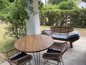 Eine Terrasse im Freien mit einem runden Holztisch, Korbstühlen und einer Holzbank mit Kissen.