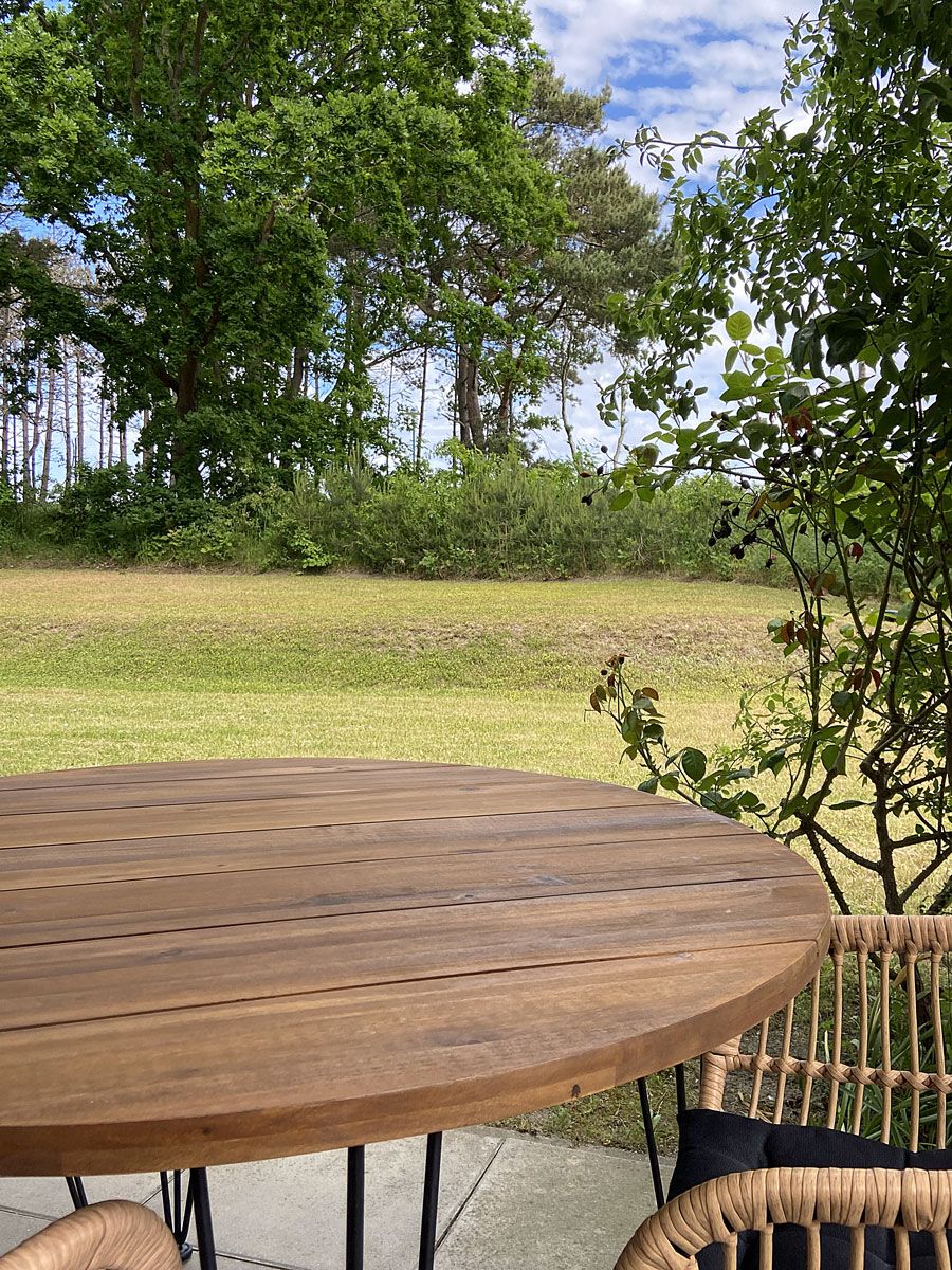 A round wooden table with a woven chair, set outdoors with a view of a grassy field and trees.