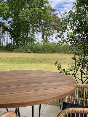 Mesa de madeira redonda com cadeira de fibra natural, disposta ao ar livre com vista para um campo gramado e árvores.