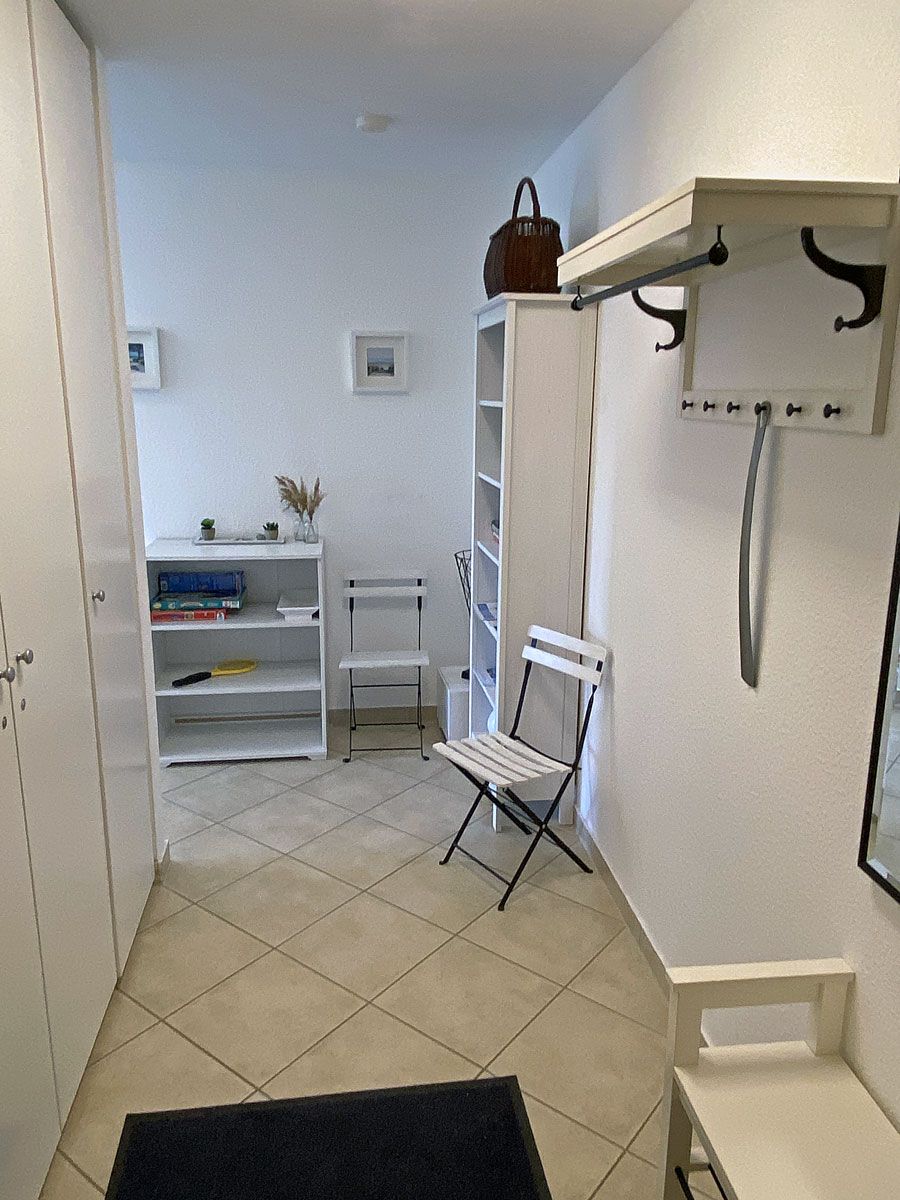 Ein Zimmer mit einem weißen Stuhl, einem Bücherregal, einem Kleiderhaken und einer Tür.