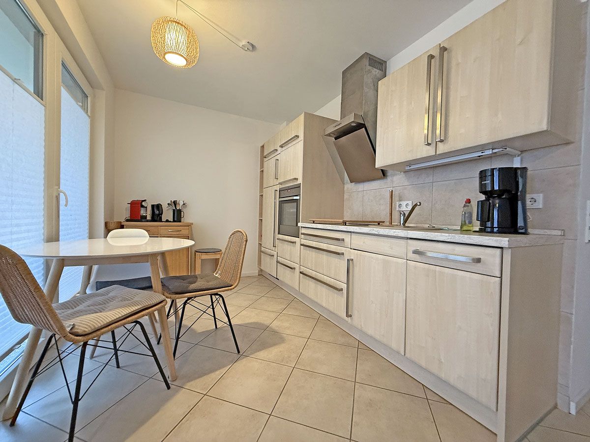 A modern kitchen with light wood cabinets, a built-in oven, and a dining area with a round table and wicker chairs.