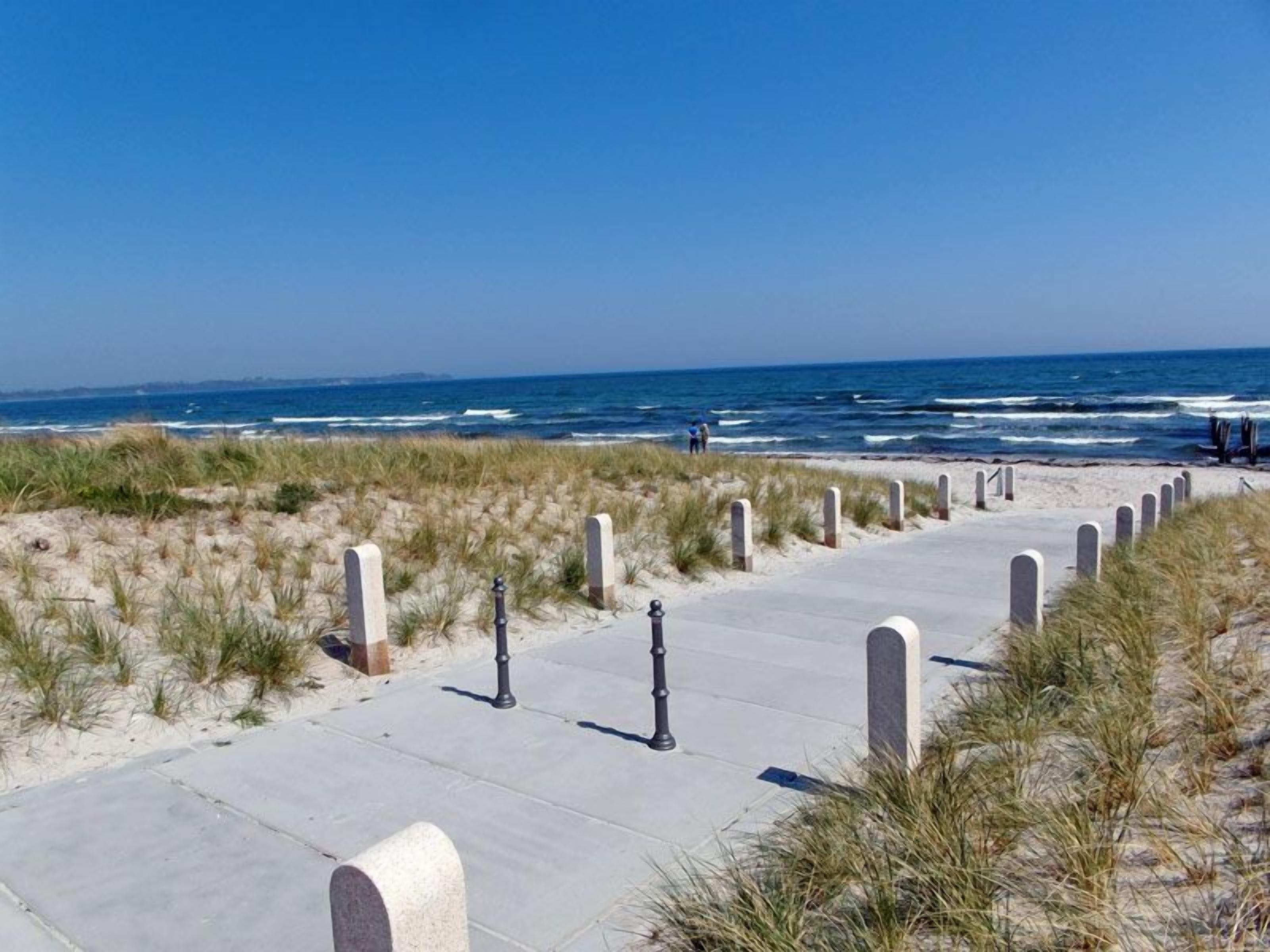 A concrete pathway leading to the beach with sand dunes and a clear blue sky.