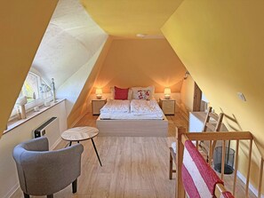 Ein Schlafzimmer mit schrägen Wänden, einem Bett mit weißer Bettwäsche, zwei Nachttischlampen, einem kleinen runden Tisch und einem Sessel.