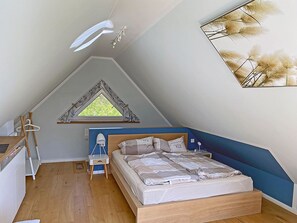 Ein Schlafzimmer mit schrägen Wänden, einem Bett mit blauer Kopfteil, einem kleinen Schreibtisch und einem Fenster mit Blick auf Grünflächen.