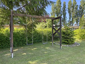 Ein hölzernes Schaukelgestell in einer grünen Wiese, im Hintergrund ein Heckenstreifen und Bäume.