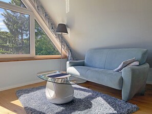 Ein modernes Wohnzimmer mit einem Glastisch, einem hellblauen Sofa und Blick auf Bäume durch ein großes Fenster.