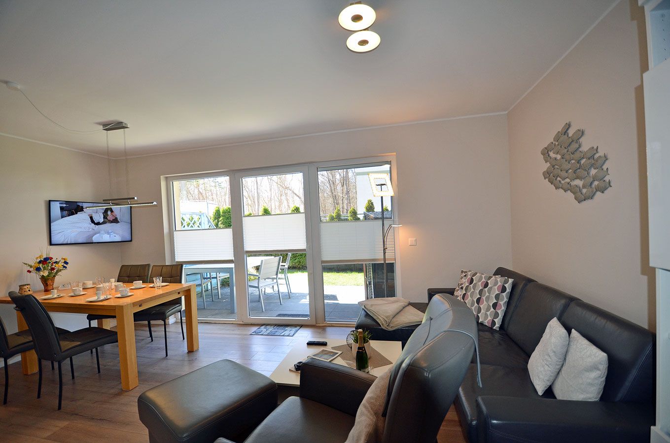 Ein modernes Wohnzimmer mit einer Couch, einem Esstisch mit Stühlen, einem an der Wand montierten Fernseher und einer großen Schiebeglastür, die zu einem Außenbereich führt.