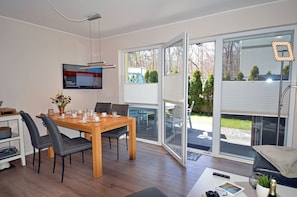 A modern dining area with a wooden table and chairs, a television mounted on the wall, and a view of the outdoors through large sliding glass doors. - First floor, modern amenities, central location, about 200 m from the sandy beach (Baltic Sea) (Juliusruh)