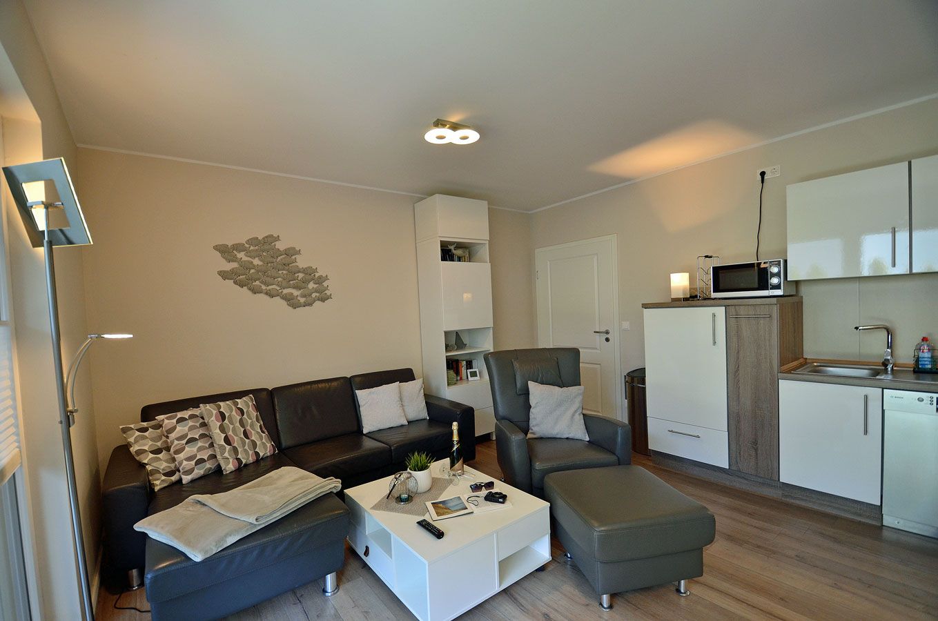 A modern living room with a sofa, armchair, ottoman, and a coffee table. There is a kitchen area with a microwave and cabinets.