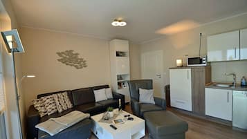 A modern living room with a sofa, armchair, ottoman, and a coffee table. There is a kitchen area with a microwave and cabinets.