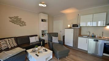 A compact living space with a sofa, armchair, coffee table, kitchenette, and a wall art piece.