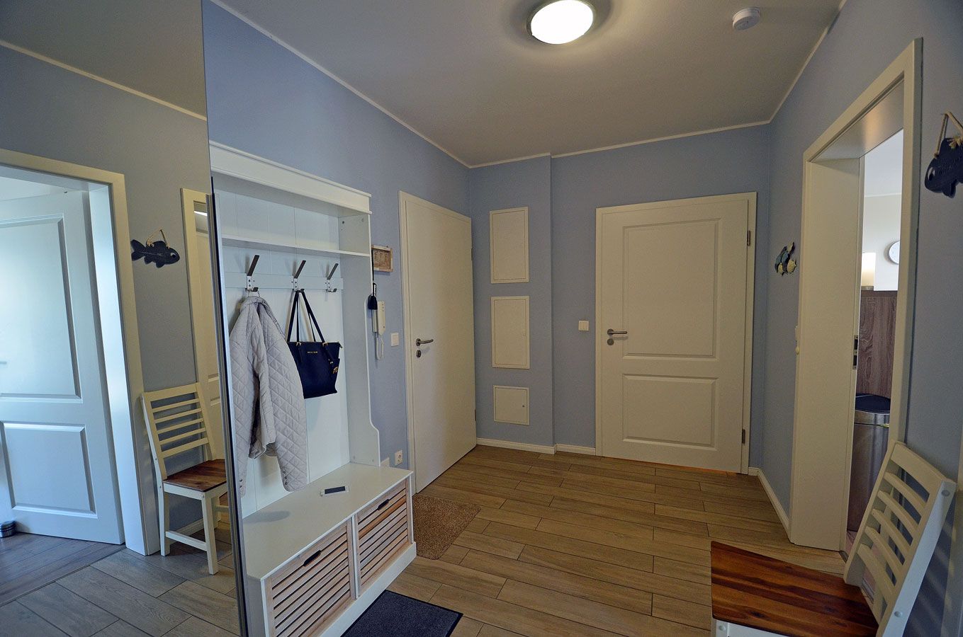 A room with a wooden floor, white walls, and a door with a mirror. There is a bench and a coat rack with a jacket and a bag.