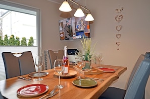 Un comedor con una mesa de madera dispuesta para cuatro, con copas de vino, platos y cubiertos. Hay sillas alrededor de la mesa y una planta en maceta sobre la mesa.
