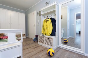 Una habitación con piso de madera, un banco blanco, un banco con espacio de almacenamiento, un perchero con chaquetas, un espejo que refleja la habitación y un balón de voleibol.