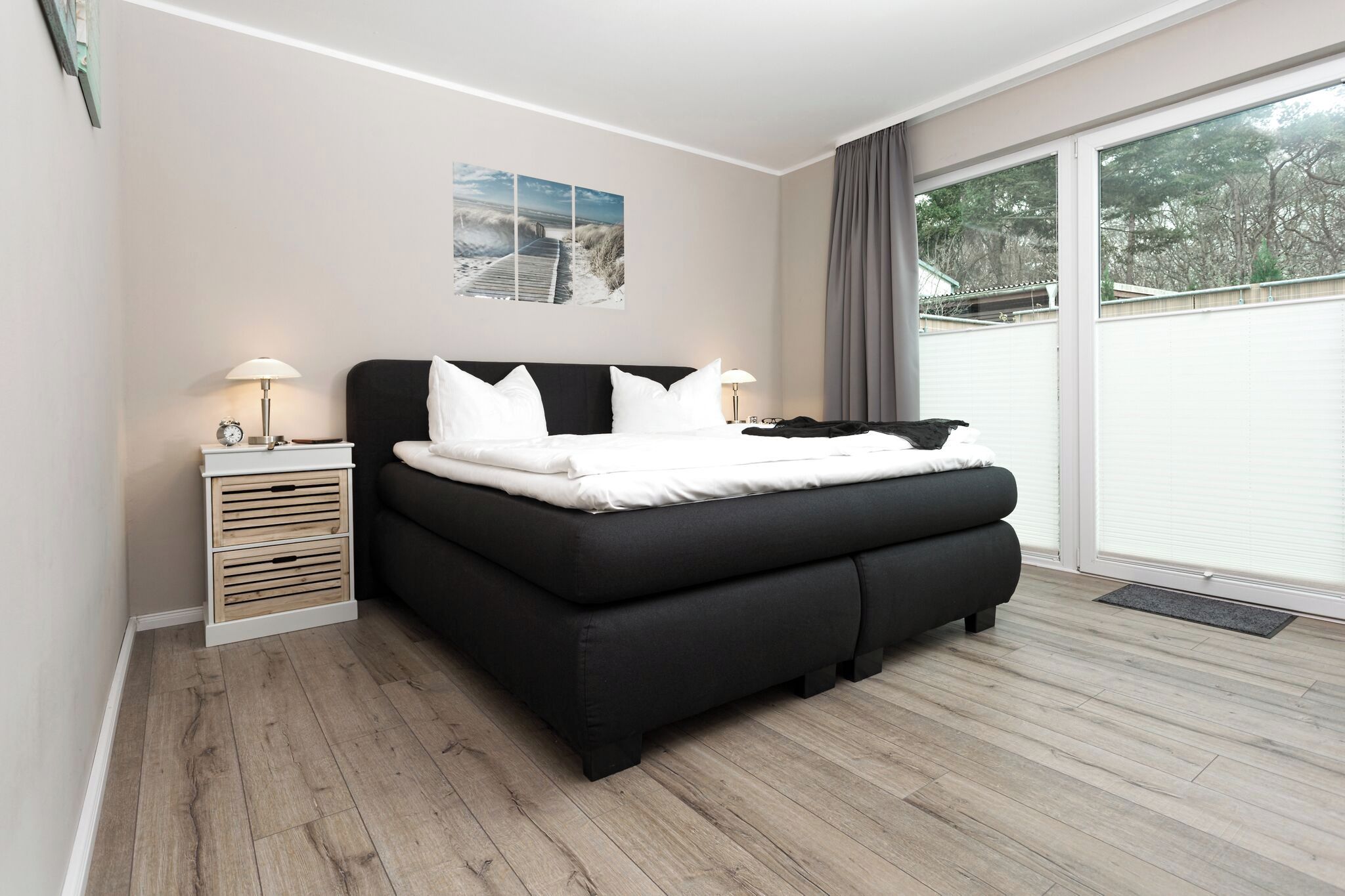 Un dormitorio moderno con un sofá cama negro, una mesita de noche con lámpara y un gran ventanal con vistas a árboles.