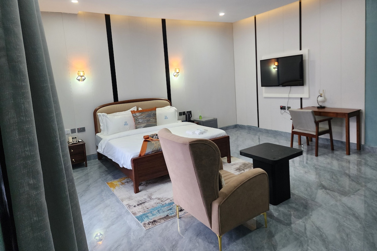 Habitación de hotel con cama, mesita de noche, silla, escritorio, televisor y una pequeña mesa con lámpara.
