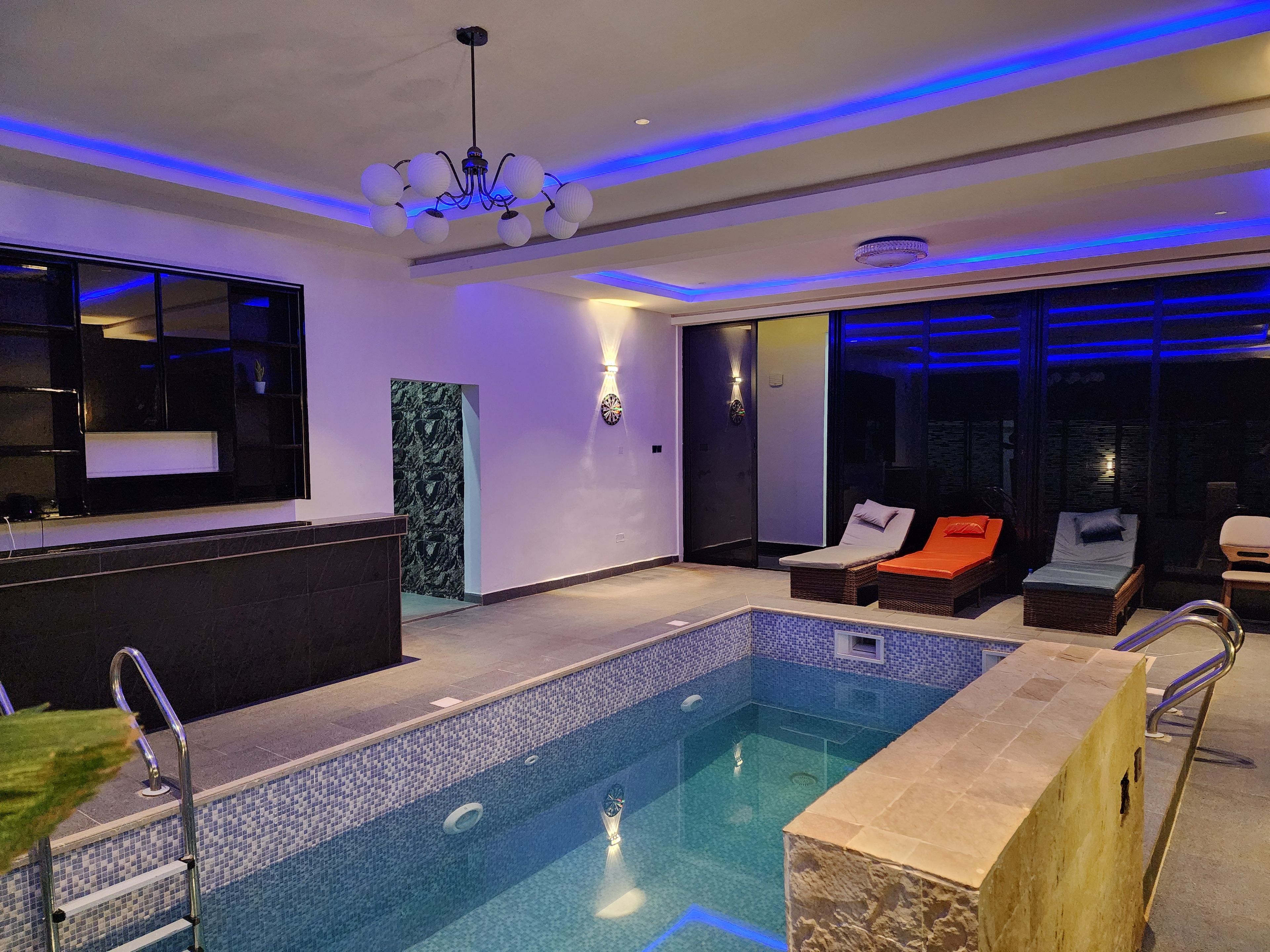 A modern indoor pool area with a clear pool, lounge chairs, and a bar area.