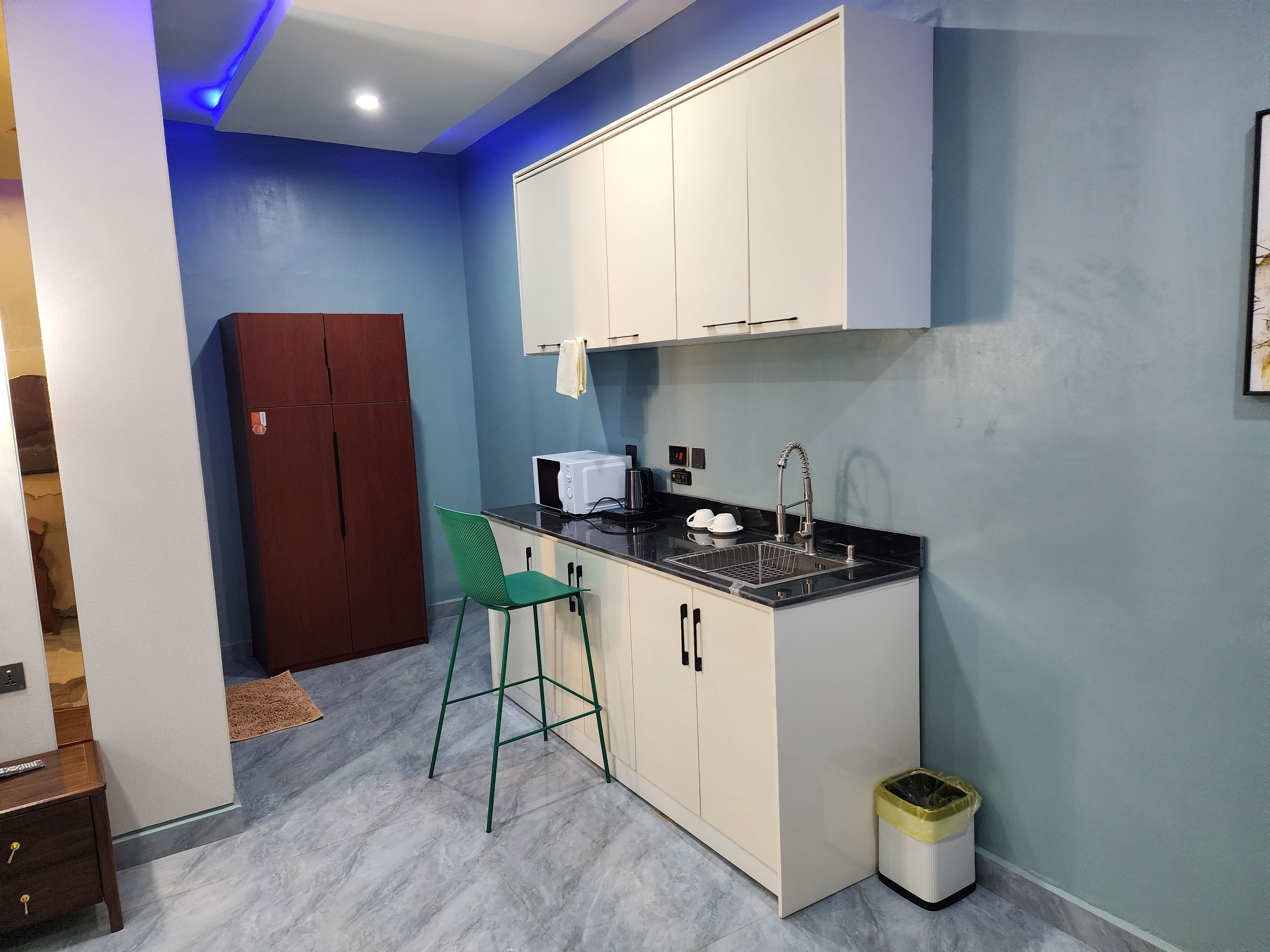 A compact kitchen with white cabinets, a black countertop, a green chair, and a small trash bin.