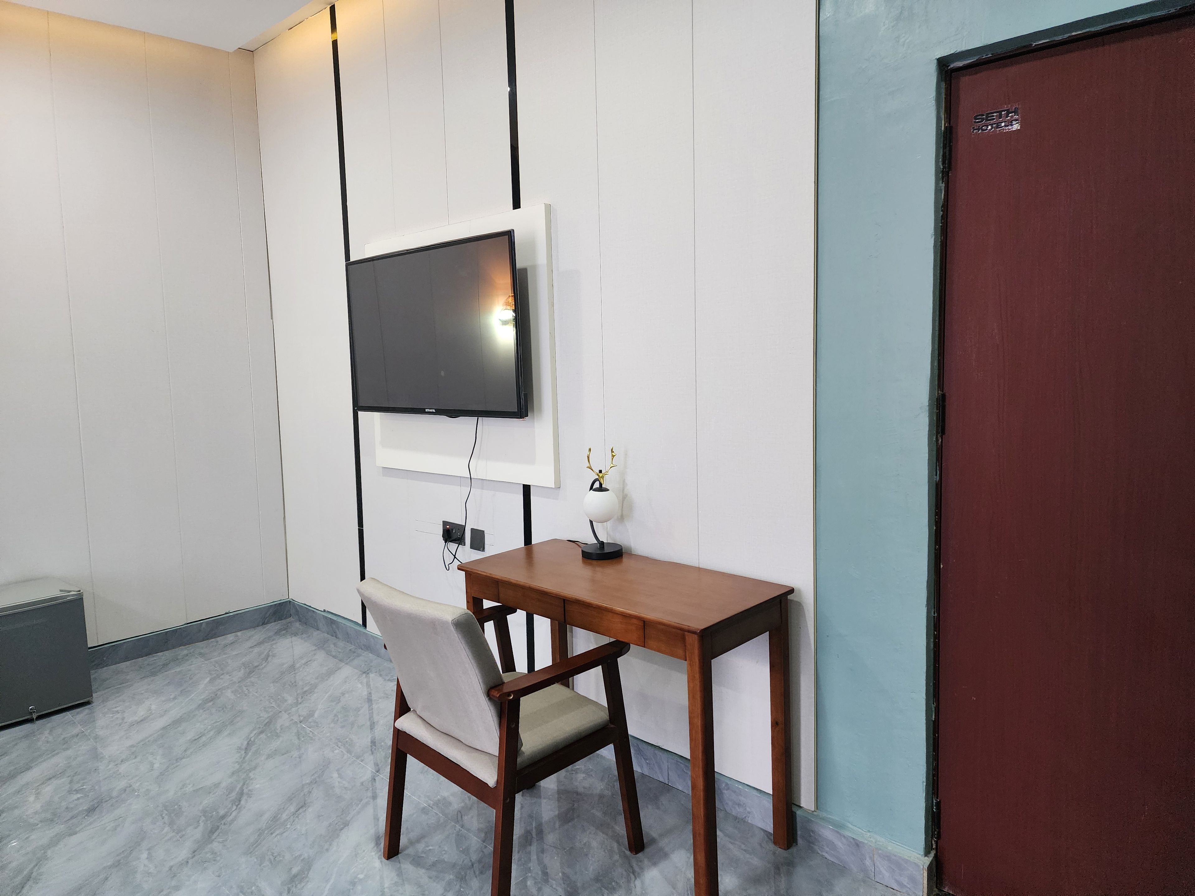A hotel room with a flat-screen TV mounted on the wall, a wooden desk with a chair, and a red door.