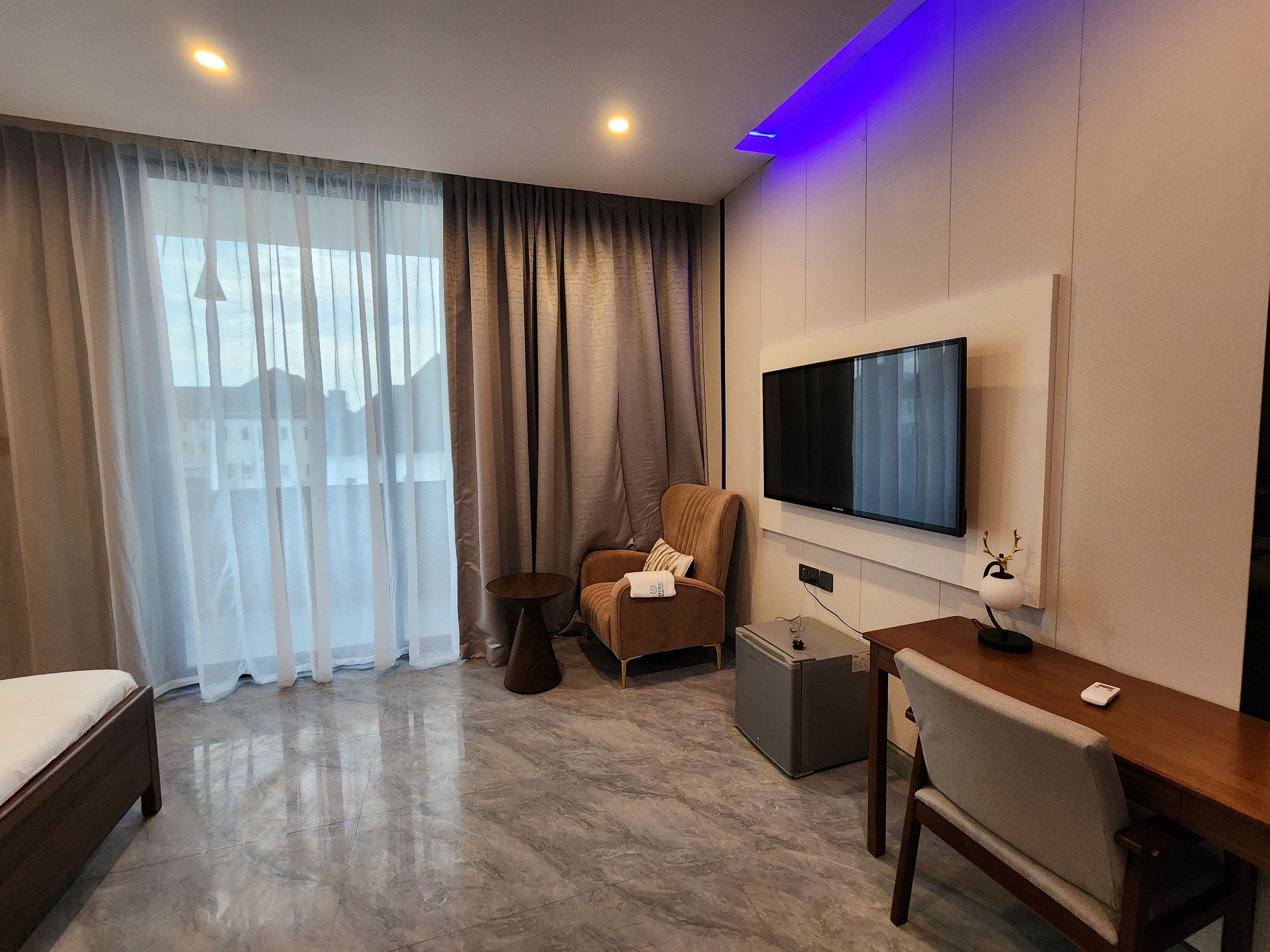 A modern hotel room with a bed, a seating area with a chair and a table, a television mounted on the wall, and a large window with sheer curtains.