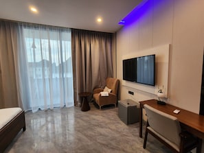 A modern hotel room with a bed, a seating area with a chair and a table, a television mounted on the wall, and a large window with sheer curtains. - Sethhotel Apartment & Suite (Asaba)