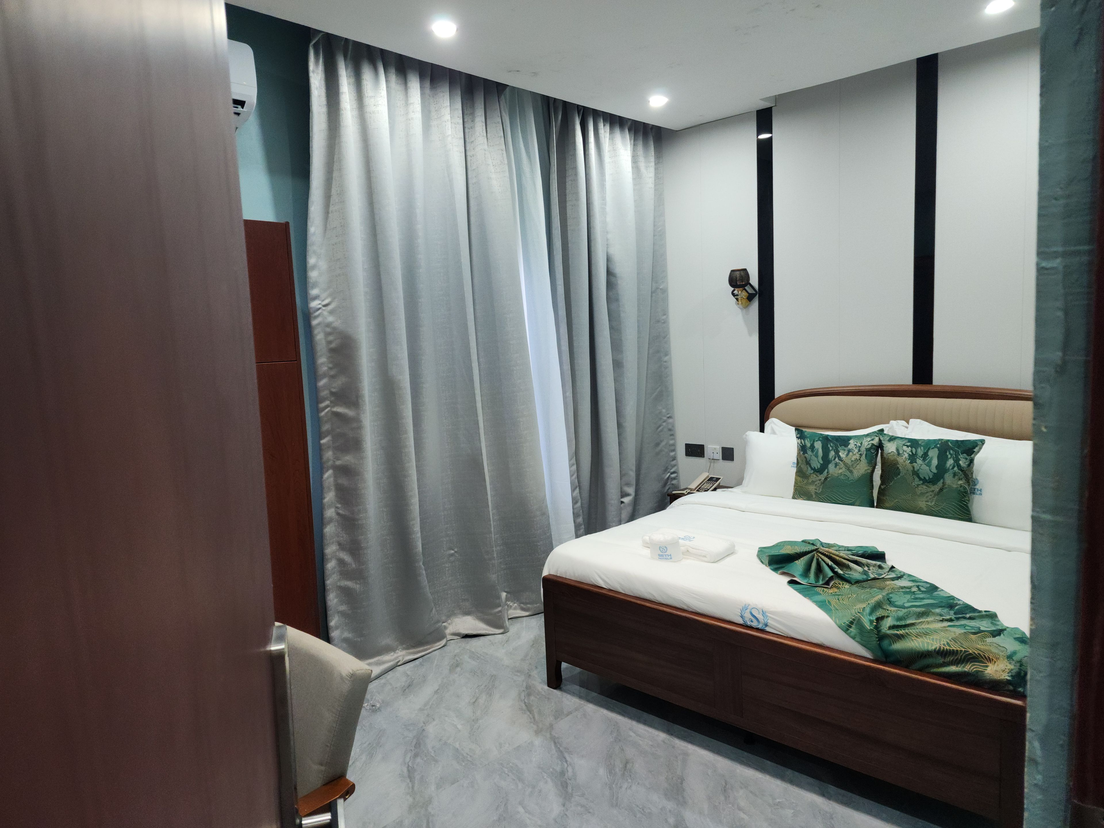 Ein Hotelzimmer mit Bett, Vorhängen, einem Nachttisch, einer Lampe und einem an der Wand befestigten Telefon.