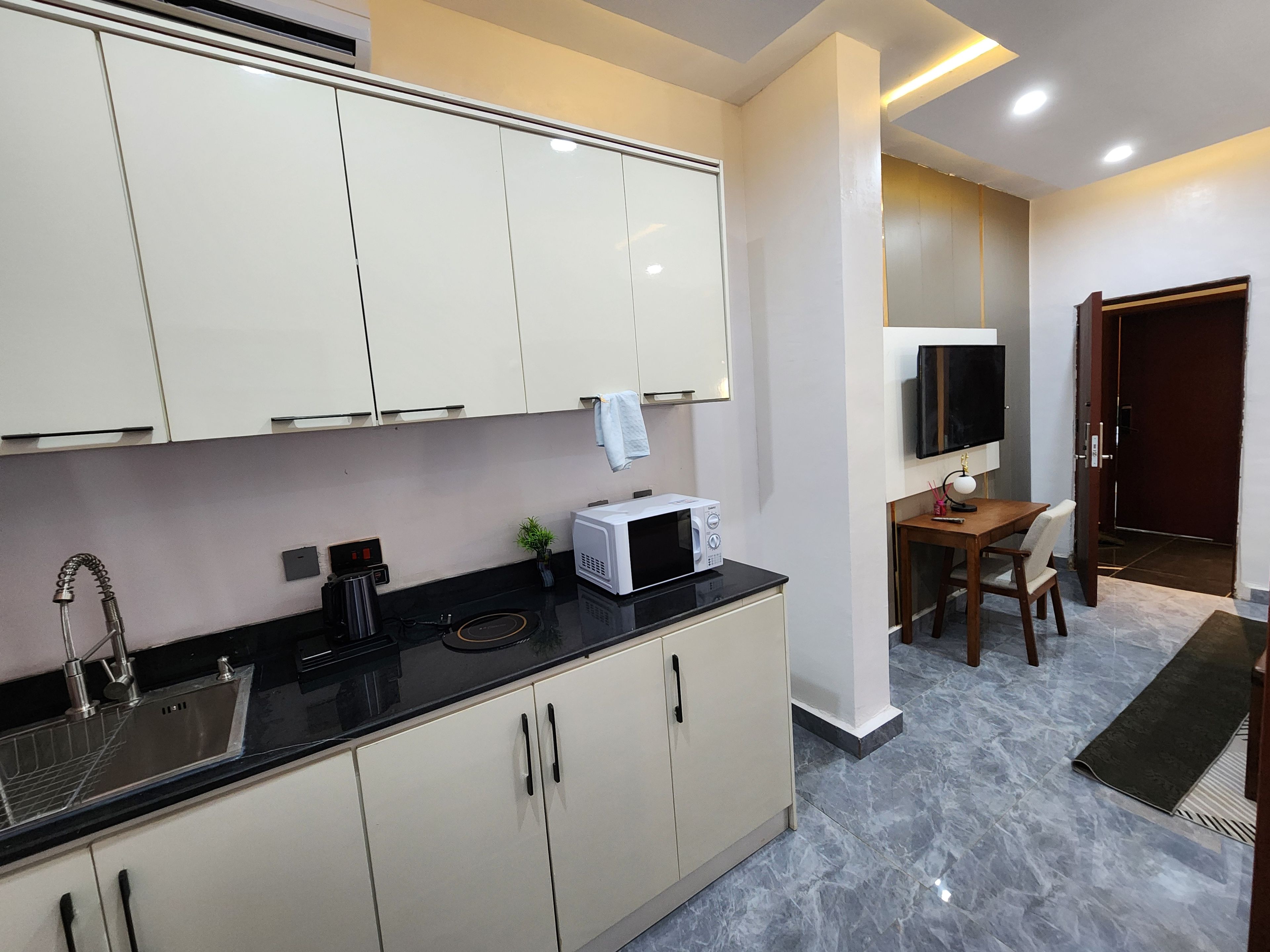 Una cocina moderna con armarios blancos, una encimera negra y un microondas. Hay una zona de comedor con una mesa y sillas, y un televisor montado en la pared.