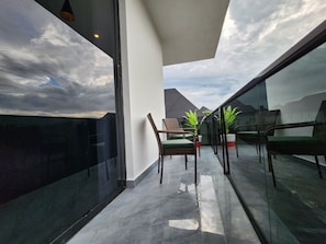 A balcony with a view of the sky and buildings, furnished with wicker chairs and a potted plant.