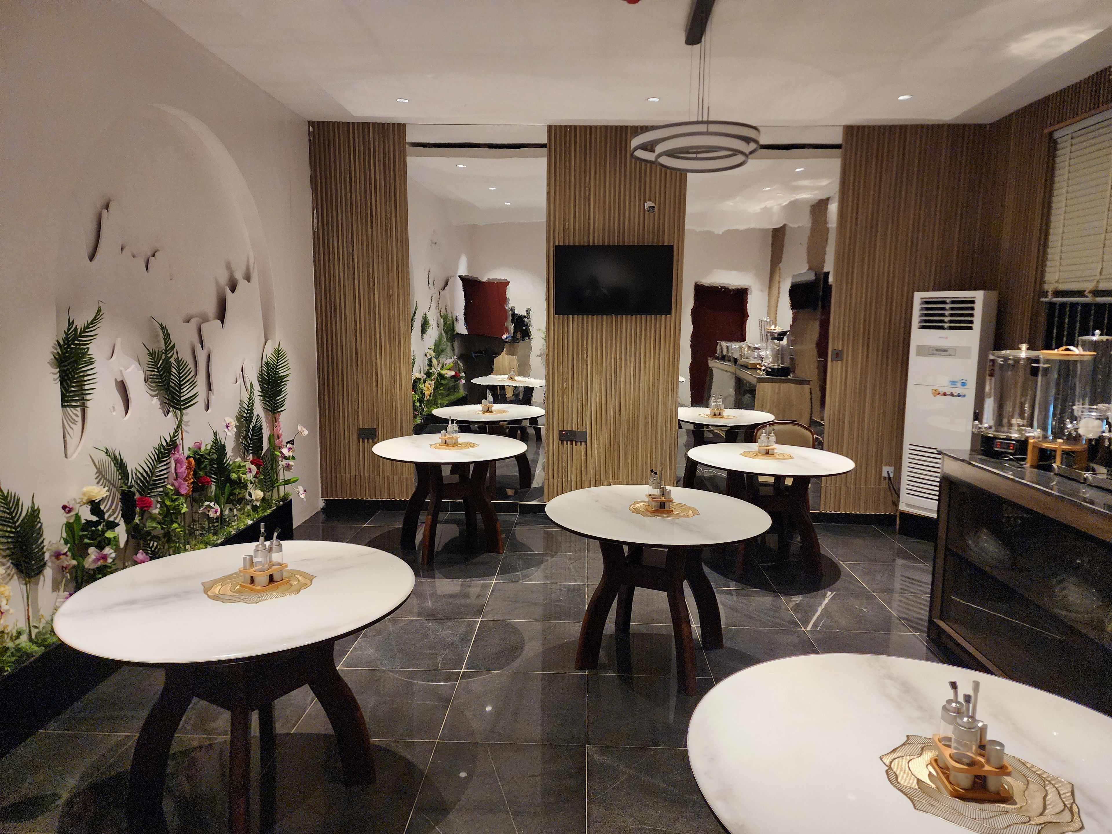 A modern interior with round tables, a flat-screen TV, and a kitchen area with a coffee machine.