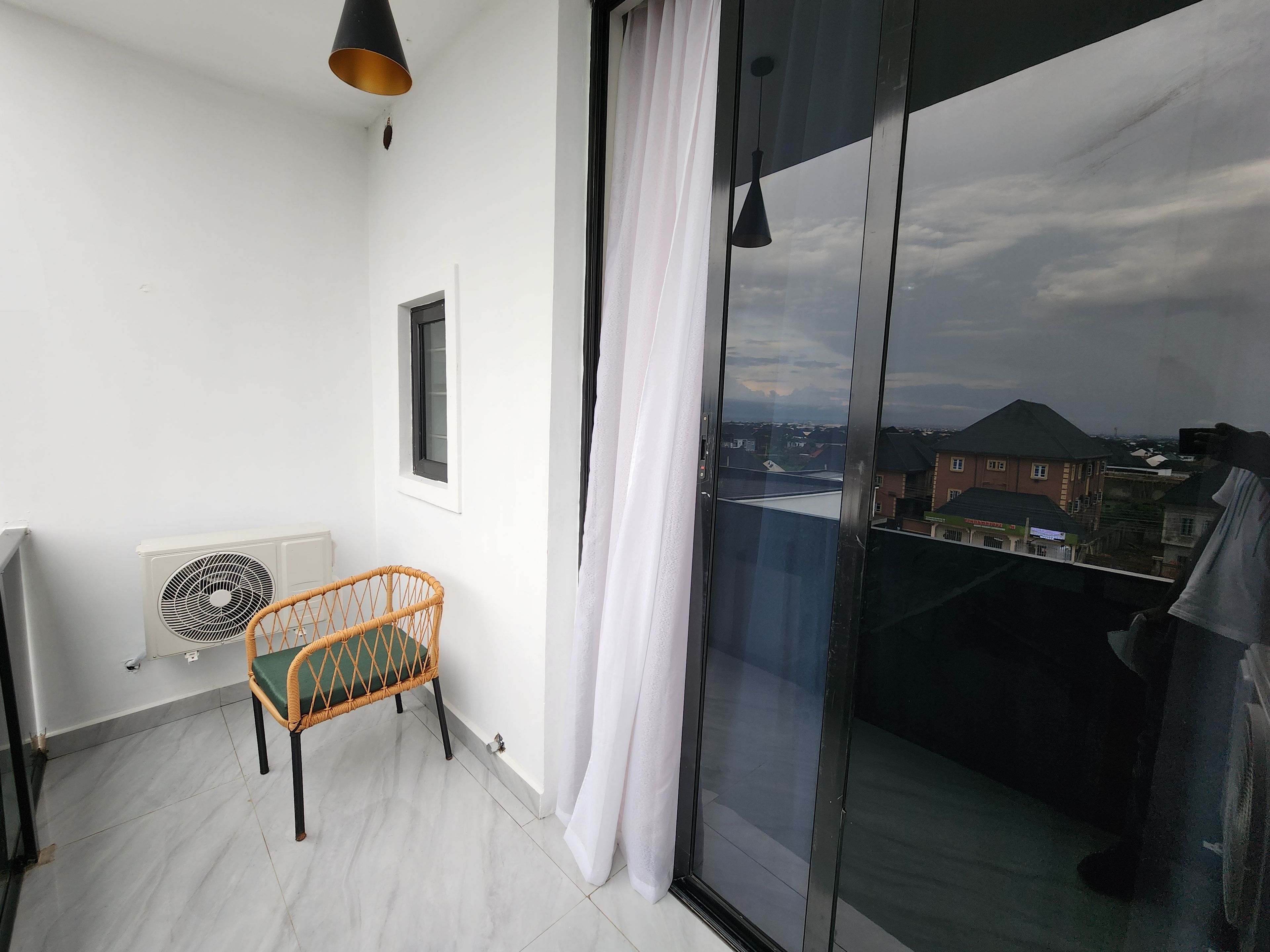 Un balcón con una silla de mimbre, una cortina blanca y vistas a edificios.