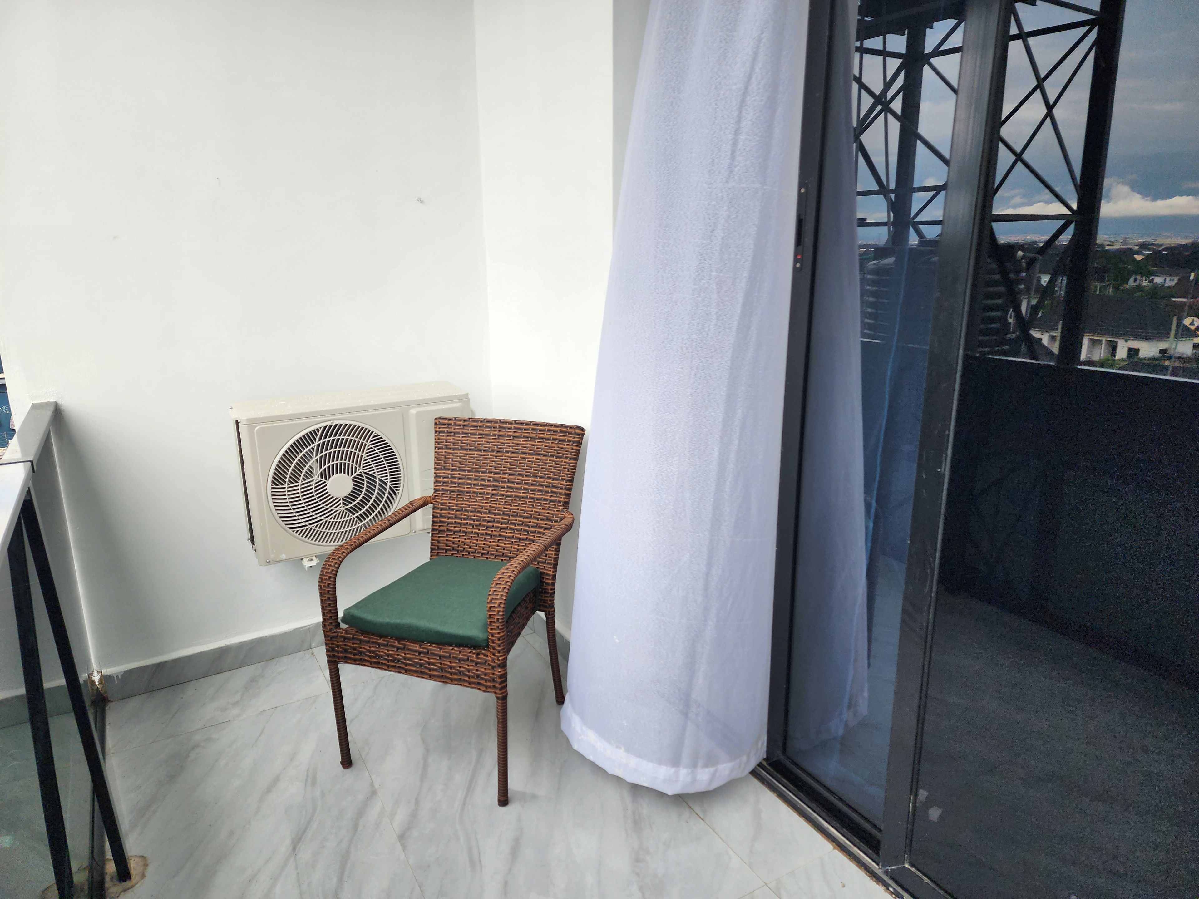 Un balcón con una silla de mimbre, una cortina blanca y vista a un edificio a través de una puerta de cristal.