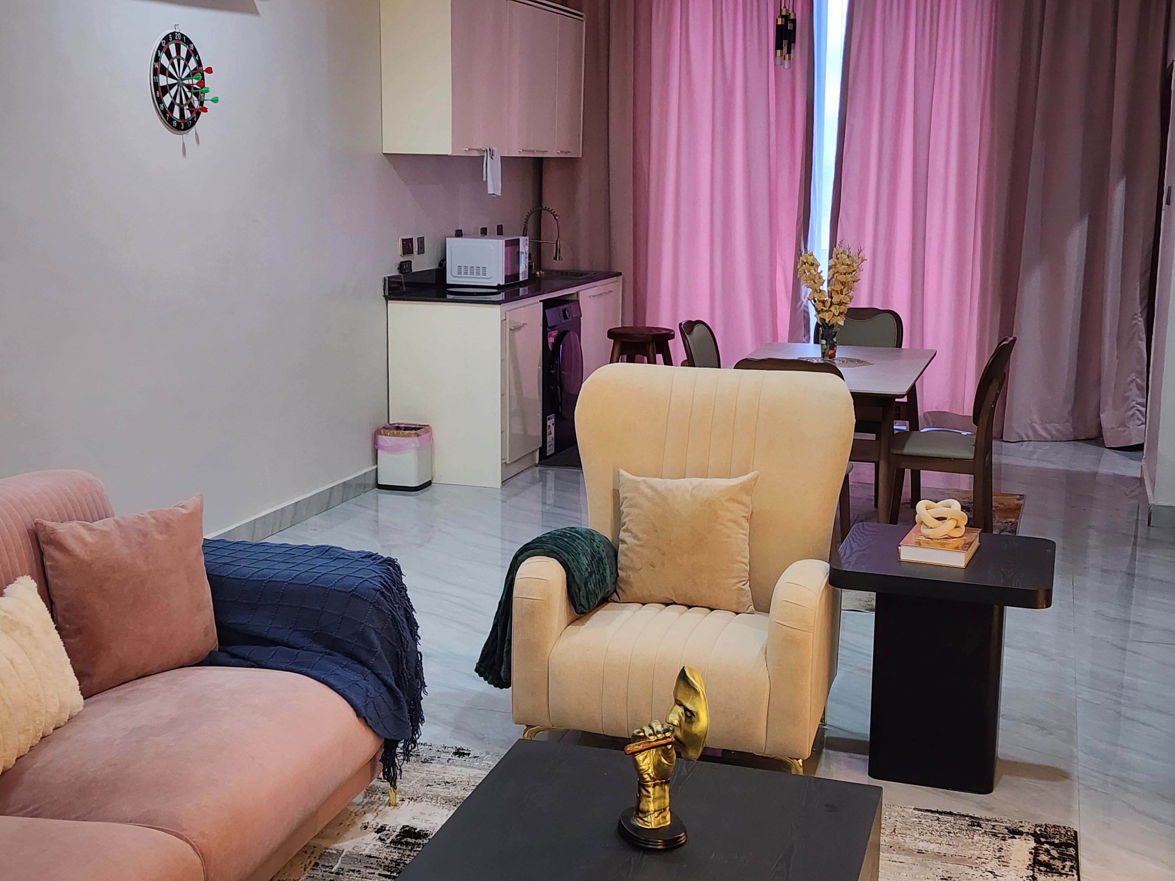 A living room with a sofa, armchair, and a coffee table with a decorative item. There is a dining area with a table and chairs, and a kitchen area with a microwave and cabinets.