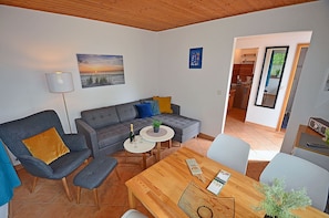 En moderne stue med en grå sofa, et træspisebord og et køkken, der kan ses gennem en åben dør.