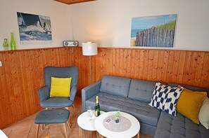 Ett rum med en grå soffa, en tillhörande fåtölj, ett litet runt bord med en flaska och ett glas, och två radioapparater som är monterade på väggen.