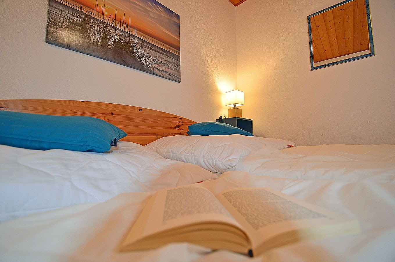 Una cama con un libro abierto, una mesita de noche con una lámpara y un cuadro en la pared.