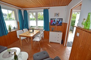 Ein Zimmer mit Holzboden, einem Esstisch mit Stühlen, einem Fernseher an der Wand und einer kleinen Kommode.
