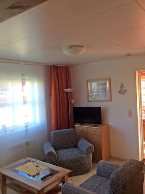 Ett litet vardagsrum med en tv, två fåtöljer, ett litet bord i trä och ett fönster med gardiner.