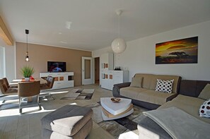 Uma sala de estar moderna com sofá, mesa de centro e televisão. Há também um espaço de jantar com cadeiras e uma mesa.