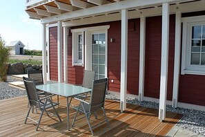 Área externa coberta com mesa e cadeiras de vidro, deck de madeira e um prédio vermelho com detalhes brancos.