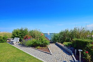 A paved pathway leading to a body of water with a sailboat in the distance, surrounded by lush greenery and a bench. - Vacation home in a very quiet location with a view of the Bodden; pets allowed; 2 bedrooms (Breege)