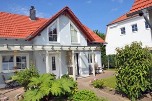 Ein zweigeschossiges Haus mit rotem Ziegeldach, einer Veranda und einem Garten mit verschiedenen Pflanzen.