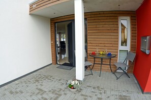 Ein kleiner Sitzbereich draußen mit Tisch und Stühlen, eine Tür zu einem Innenraum sowie ein Blumenkorb auf dem Boden.