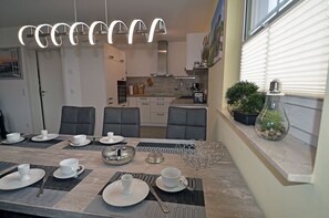 Ein Essbereich mit Tisch für vier Personen, eine Küche im Hintergrund und ein Fenster mit Jalousien.