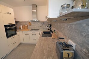 Uma cozinha moderna com armários brancos, forno embutido, exaustor de aço inoxidável, bancada de madeira e torradeira.